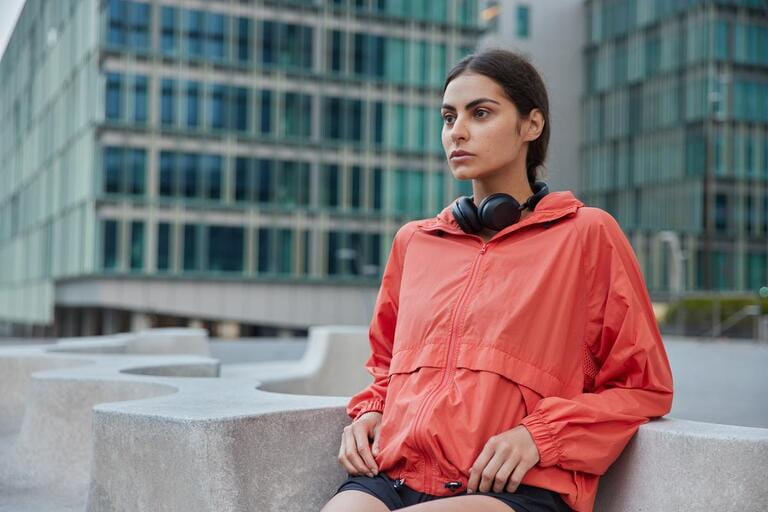 Young female in windbreaker leans with elbows on stone does exercises outdoors motivates to lead healthy lifestyle goes in for sport on city street looks forwards poses against urban building behind