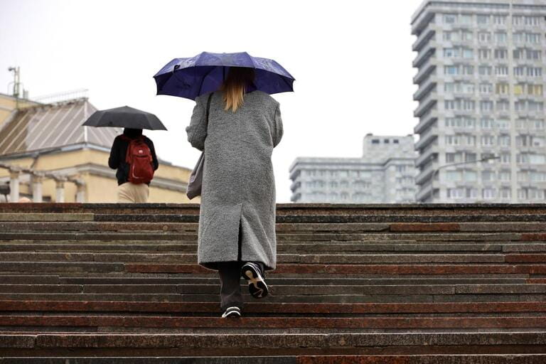 Woman in statement coat holding umbrella walking up steps on city building floor. Rain on the spring city