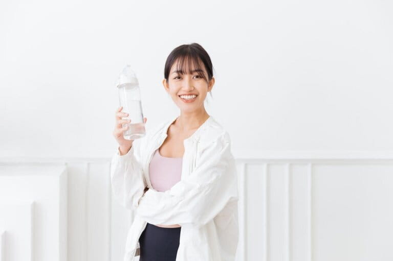 Asian woman with long black hair tied up, wearing sportswear and white sports jacket. Happy smiling in front of camera and holding a water bottle standing on white background.