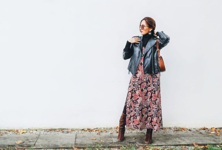 Smiling woman dressed in vintage fashion colorful long skirt with black leather biker jacket with brown leather flap pocket posing on white wall background.