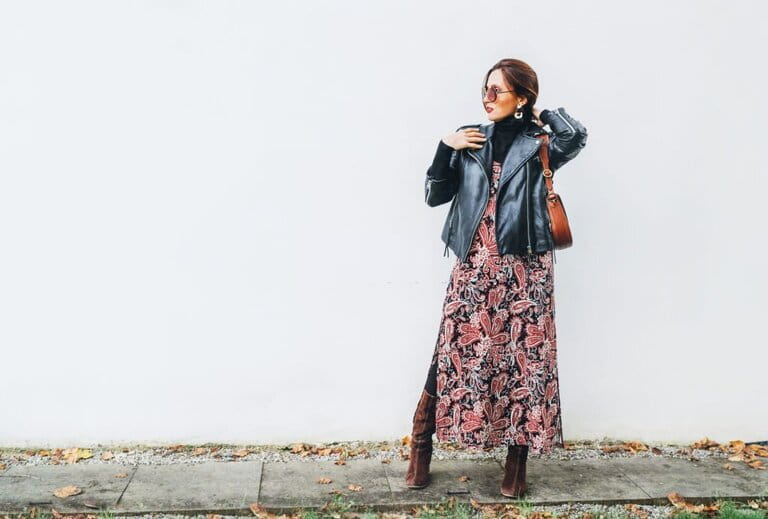 Smiling woman wearing colorful boho fashion long skirt with black leather biker jacket with brown leather flap pockets posing on white wall background. Smiling woman wearing colorful boho fashion long skirt with black leather biker jacket with brown leather flap pockets posing on white wall background.